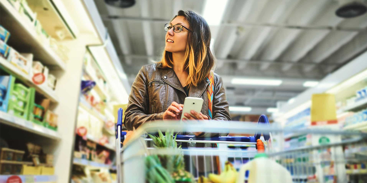 Woman shopping in a grocery store with a phone and cart.